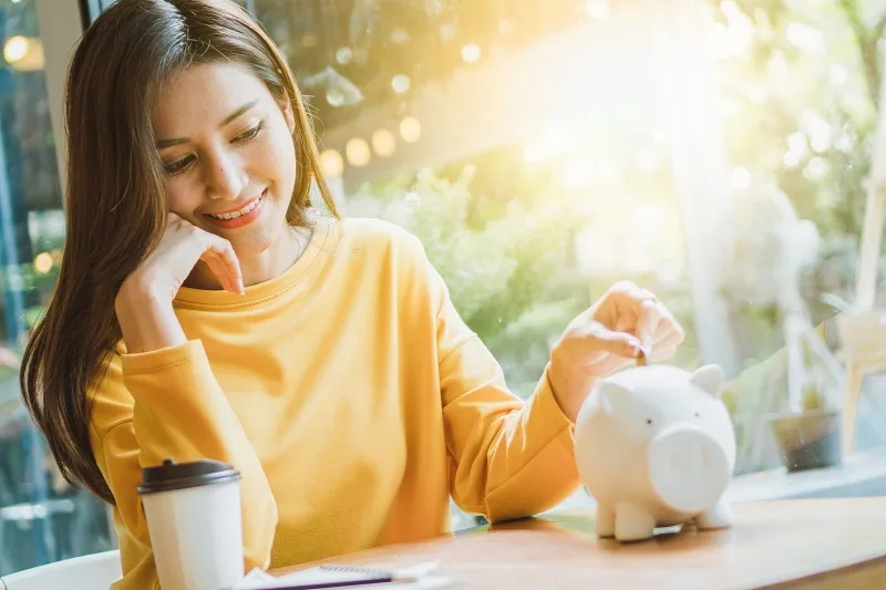 黄色のセーターを着た女性が、貯金箱に硬貨を入れながら微笑んでいる様子(保険料の無事故割引による貯蓄イメージ)